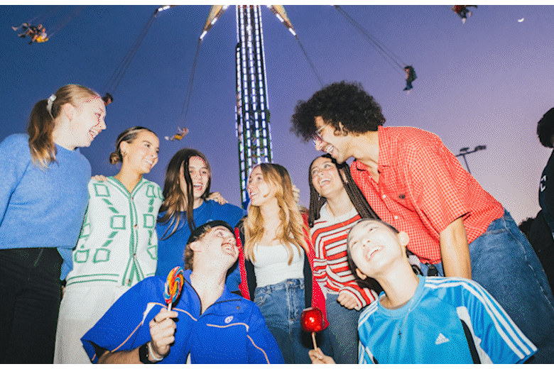 A group of young friends gathered at a funfair at dusk, laughing and smiling, with a swing carousel in the background.