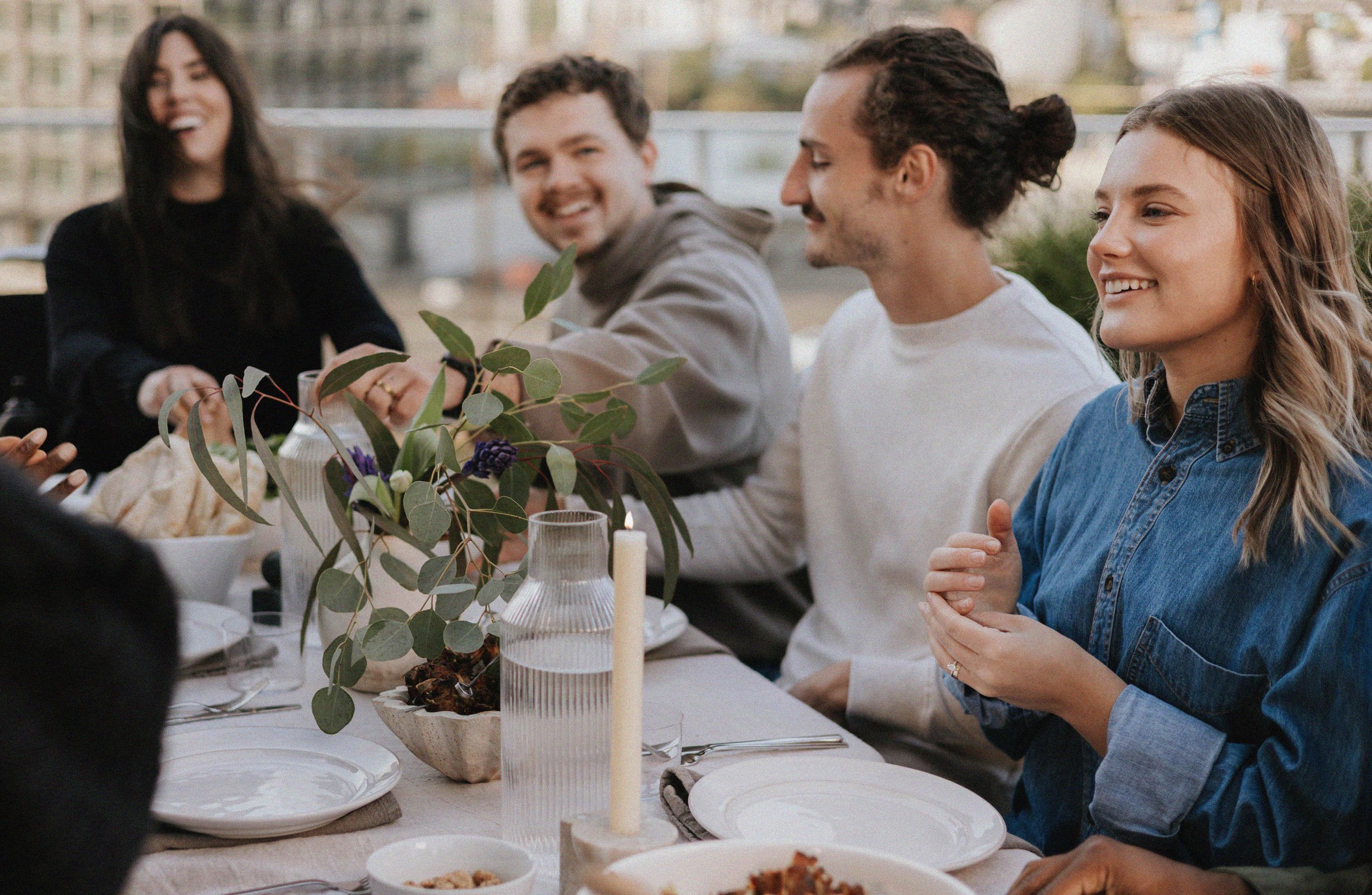 Group of five young adults sitting at a table outdoors, smiling and enjoying a meal together.
