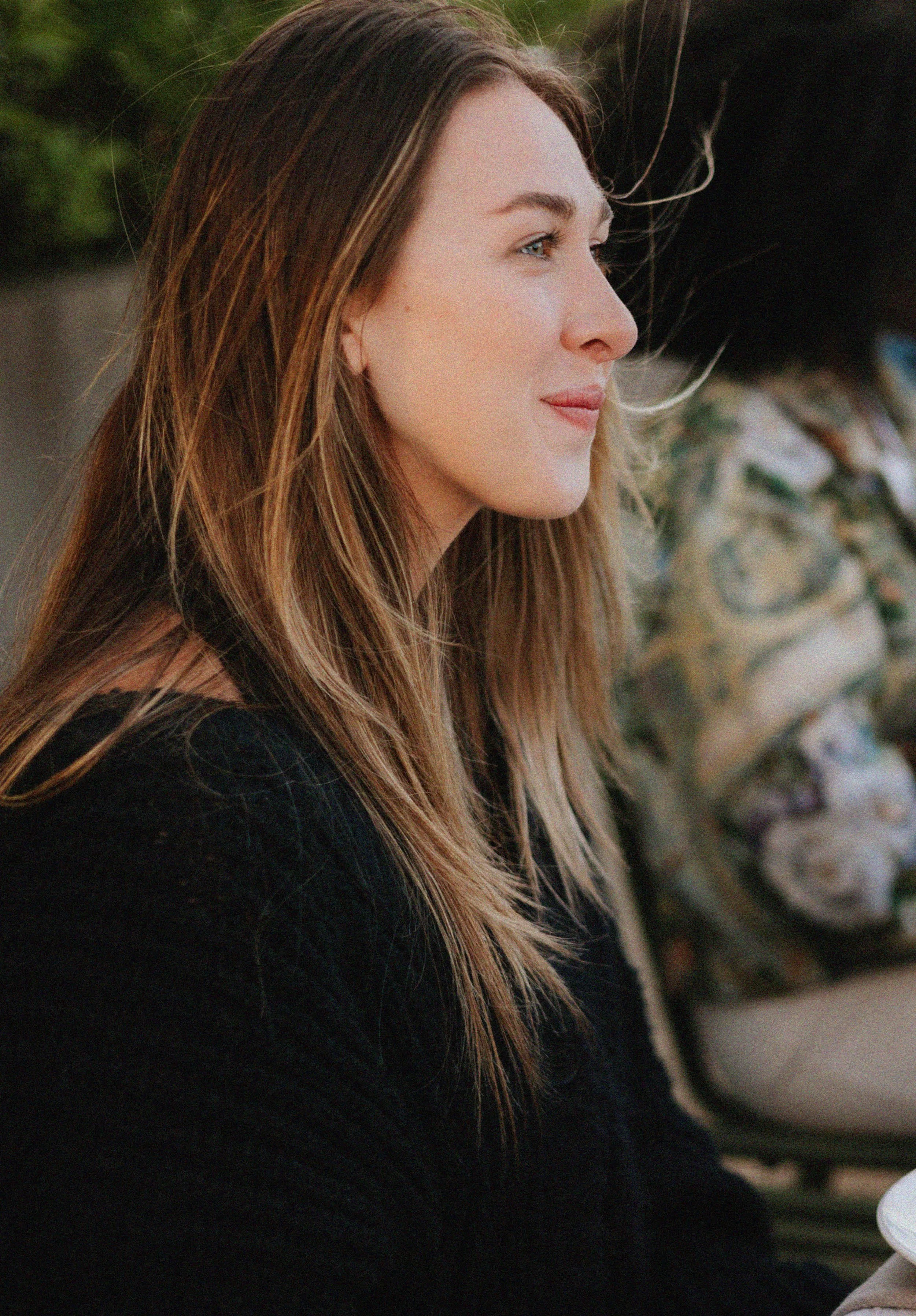 Profile of a young woman with long light brown hair and fair skin, sitting outdoors beside another person wearing a camouflage shirt.