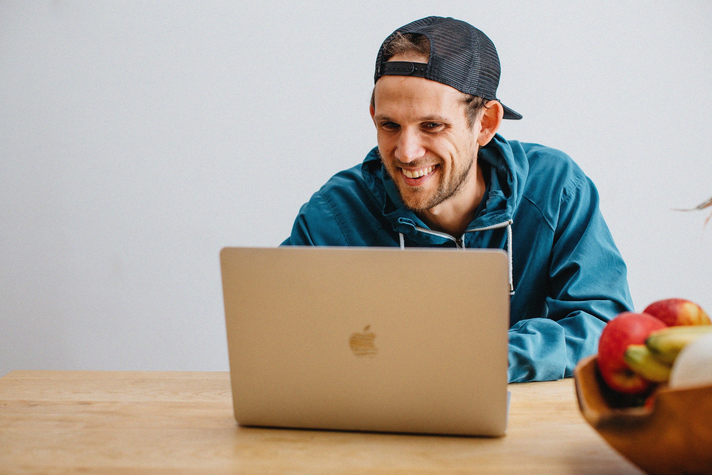 A man wearing a dark baseball cap and a blue hoodie is smiling while looking at a silver Apple laptop, sitting at a wooden table with a bowl of fruit on the side.