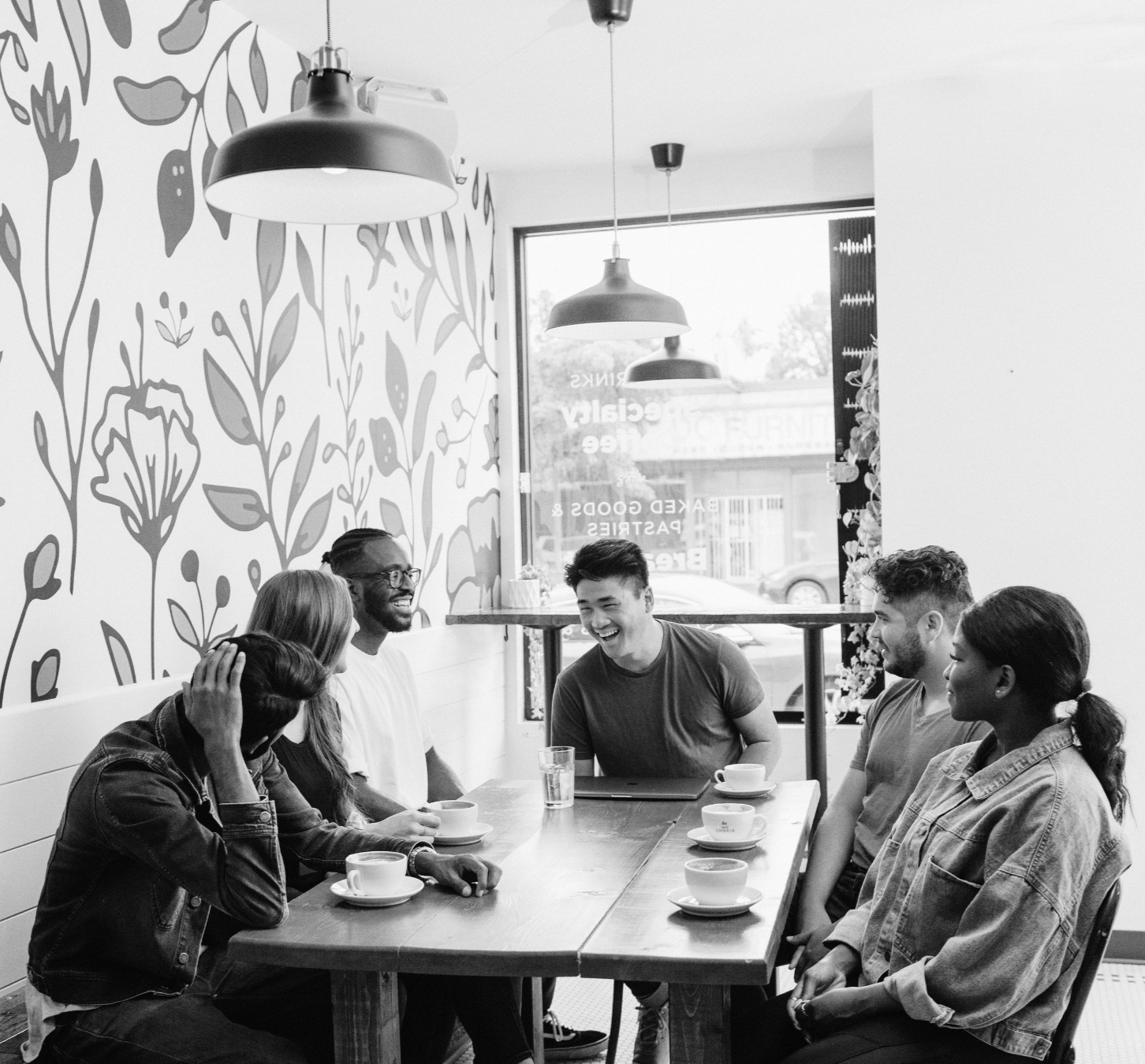 A group of five diverse young adults sitting around a wooden table in a cafe, engaging in conversation and laughing, with cups of coffee or tea in front of them, natural light coming through large windows, modern decor including a floral wallpaper behind them, and pendant lights hanging from the ceiling.